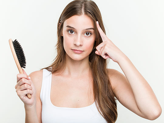 mulher branca com cabelos castanhos segurando uma escova com a mão direita e com a mão esquerda aponta para a cabeça