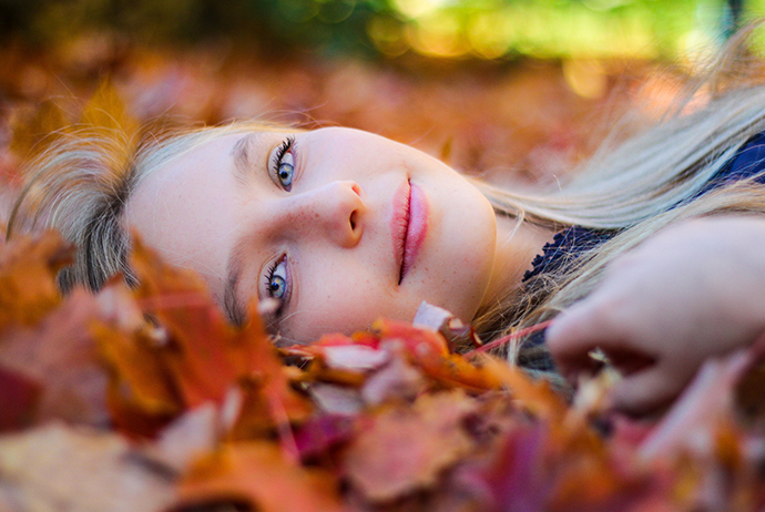 Uma mulher branca, com cabelos loiros e olhos azuis está no centro da imagem. Ela está deitada sobre folhas secas, olha para a frente e sorri. Ela veste uma blusa azul.