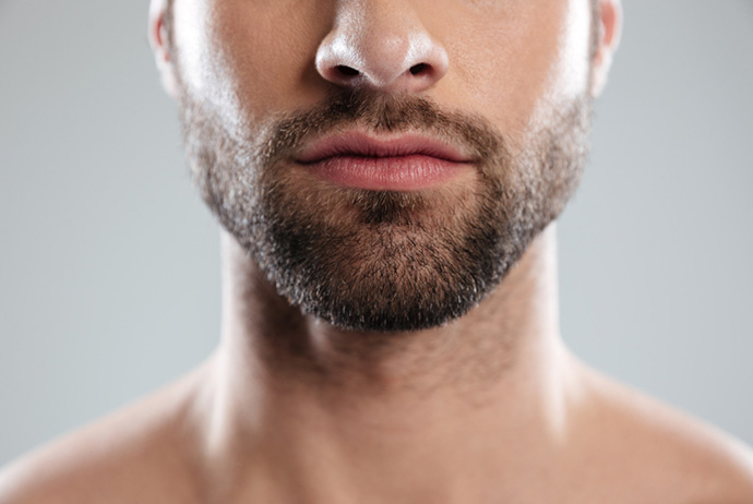 Foto em close up da metade inferior do rosto de um homem branco de barba castanha. Em seus lábios há uma seringa com agulha. O homem realiza um procedimento de preenchimento labial. O fundo da imagem é azul claro e está desfocado.